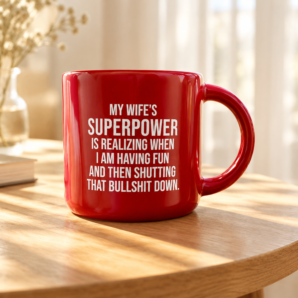 A red ceramic mug with white text reading 'MY WIFE'S SUPERPOWER IS REALIZING WHEN I AM HAVING FUN AND THEN SHUTTING THAT BULLSHIT DOWN.' sits on a wooden table in a softly lit indoor setting.