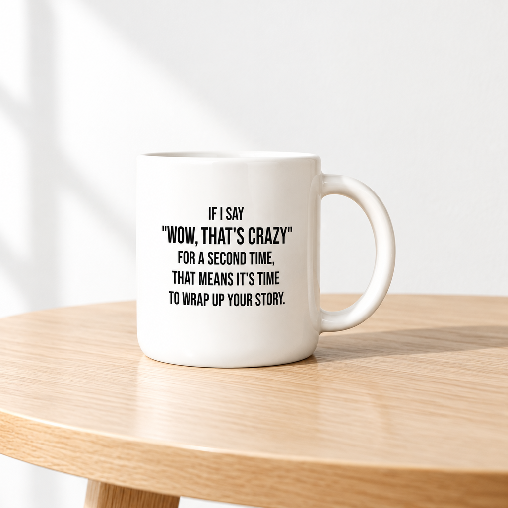 A white ceramic mug with black printed text sits on a light wood table against a white wall with soft window shadows.