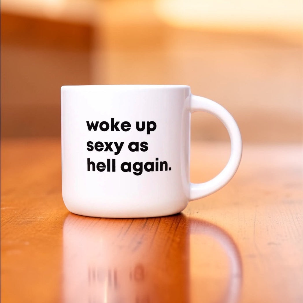 White mug with text 'woke up sexy as hell again.' on a wooden surface.