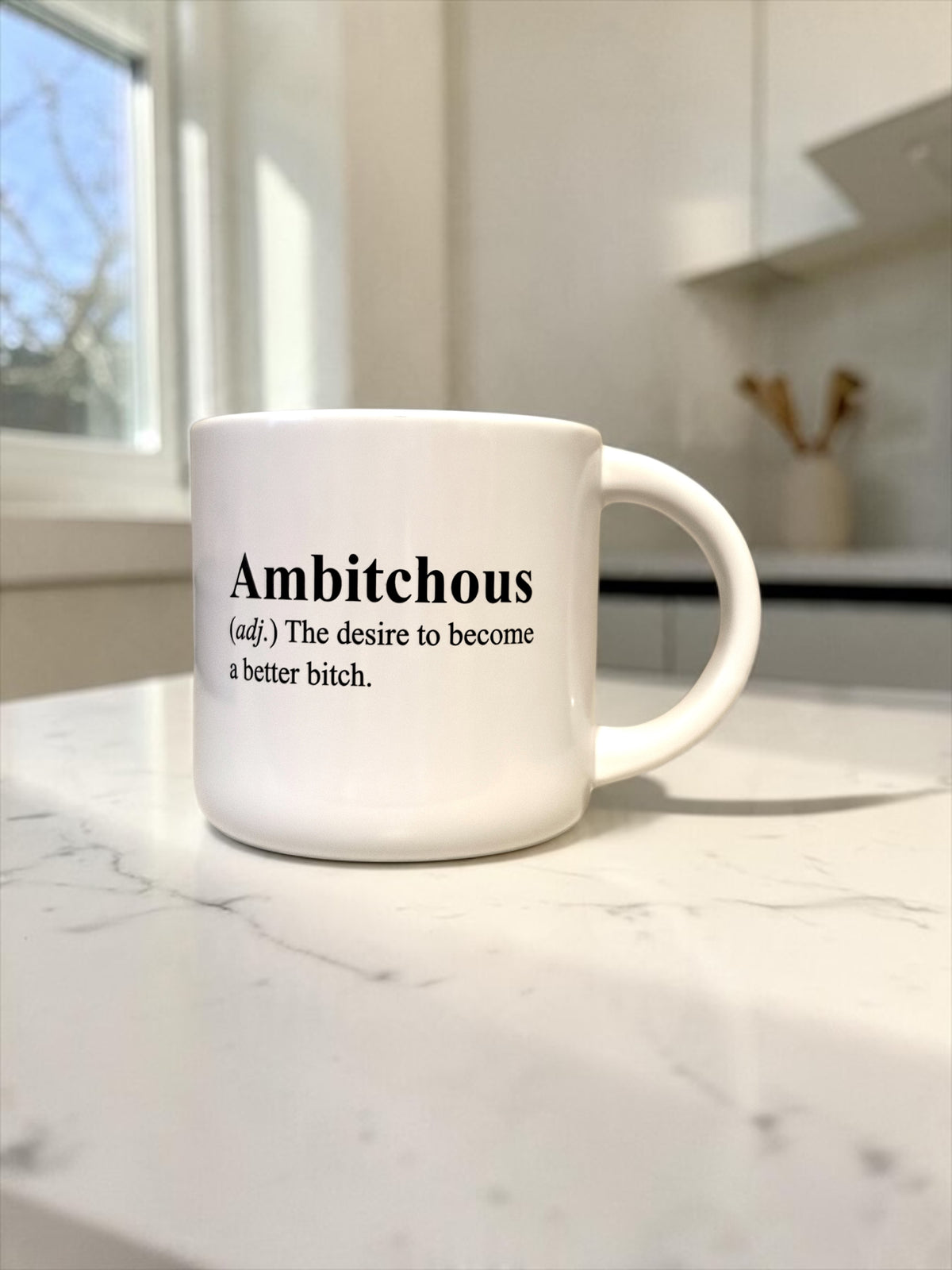 A white ceramic coffee mug with black printed text reading 'Ambitious (adj.) The desire to become a better bitch.