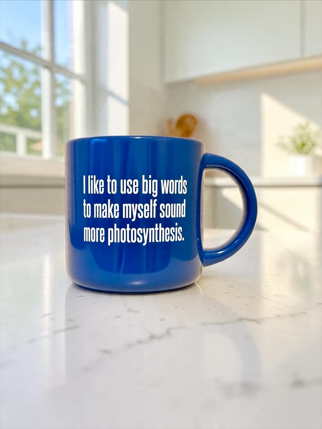 A blue ceramic mug with white printed text reading, 'I like to use big words to make myself sound more photosynthesis,' sitting on a white marble kitchen counter with a blurred kitchen background.