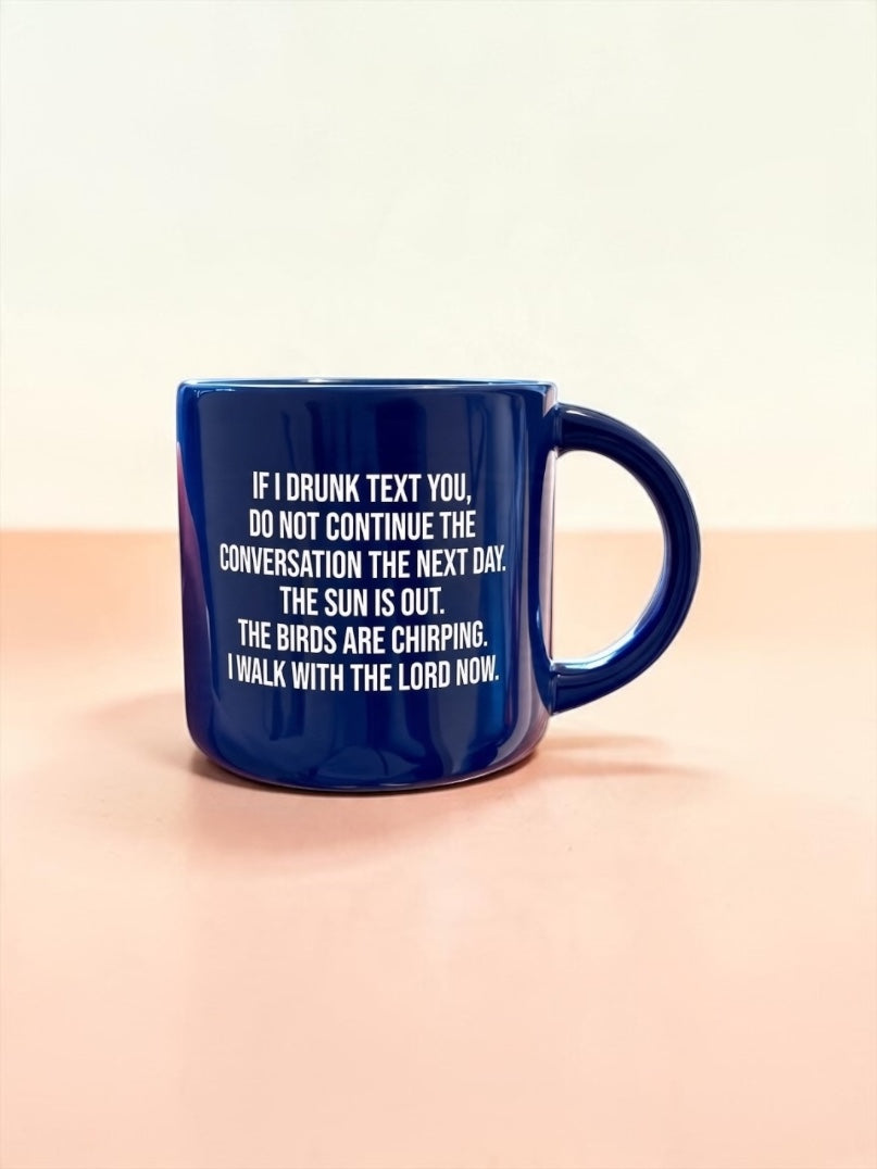 A dark blue ceramic mug with white printed text, placed on a light peach-pink surface against a cream background.