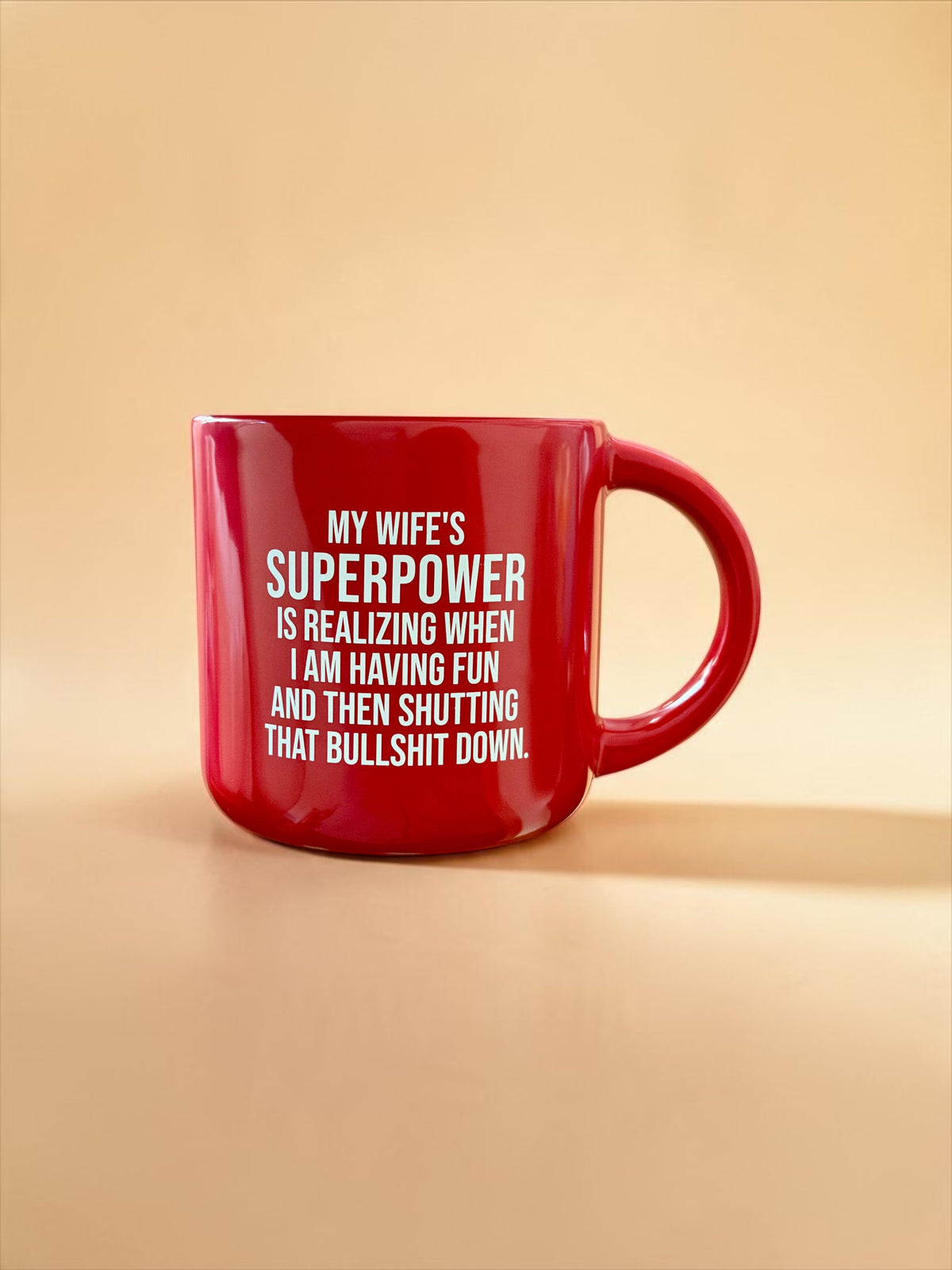 A glossy red ceramic mug with white printed text that reads, 'MY WIFE'S SUPERPOWER IS REALIZING WHEN I AM HAVING FUN AND THEN SHUTTING THAT BULLSHIT DOWN.' The mug is photographed on a smooth, solid warm beige background.