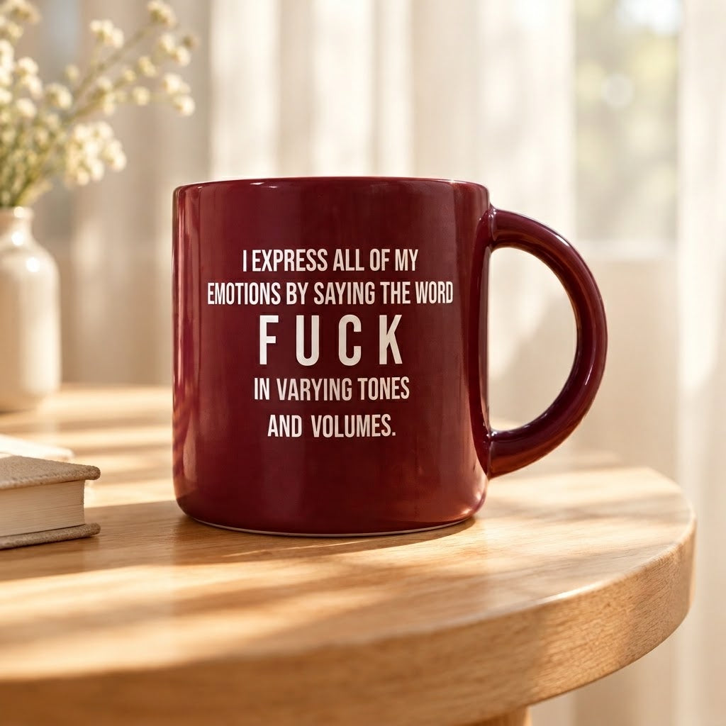 A dark red ceramic mug with white printed text sits on a round wooden table in a softly lit room. The mug reads: 'I EXPRESS ALL OF MY EMOTIONS BY SAYING THE WORD FUCK IN VARYING TONES AND VOLUMES.